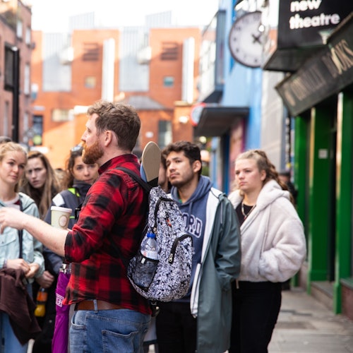 一位穿红色格子衬衫的男子在城市街道上与一群人交谈，手里拿着杯子，背着一个带滑板的背包。
