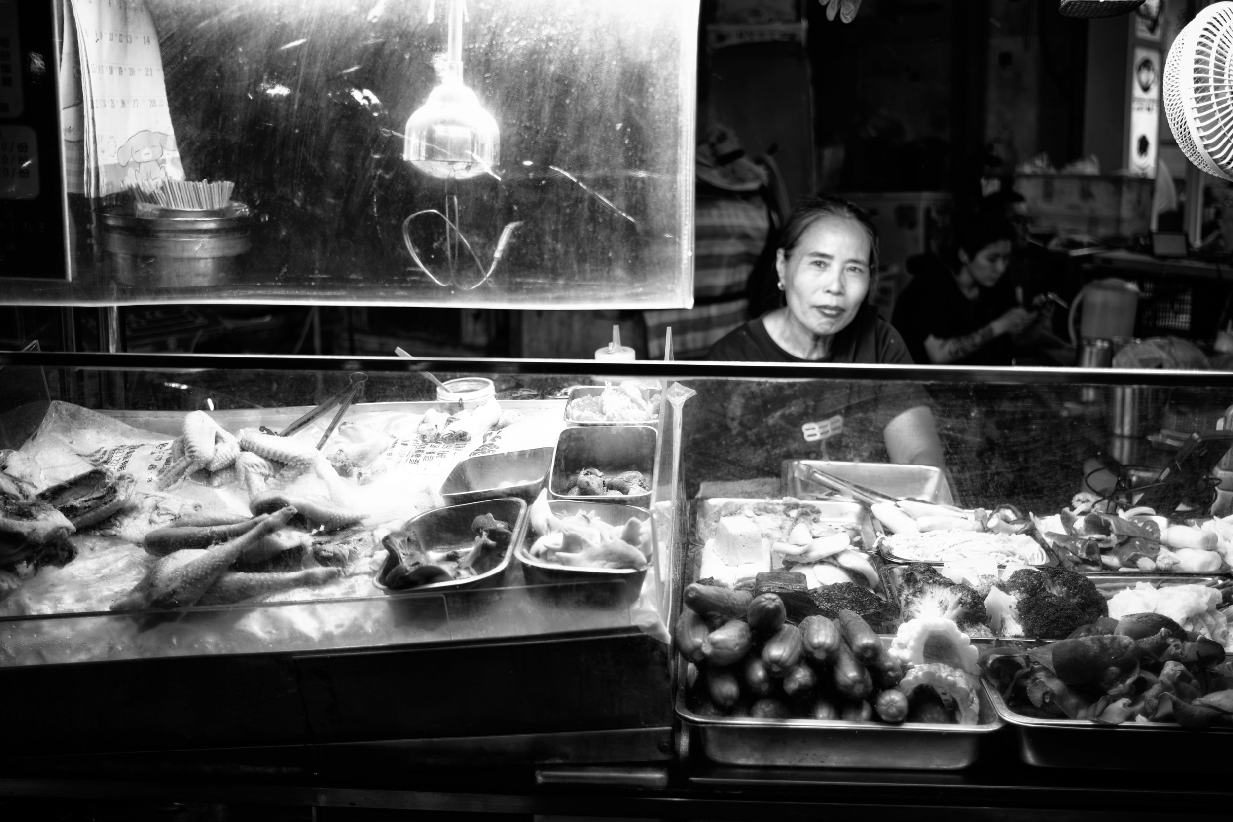 Black and white photograph of a food stall worker standing behind trays of ingredients inside a glass display case.