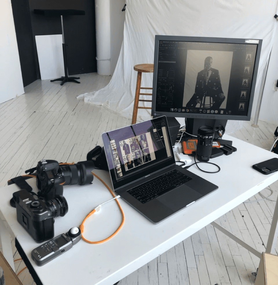 A white table with DSLR and medium-format cameras, laptop, and monitor displaying black-and-white portraits in a photo studio
