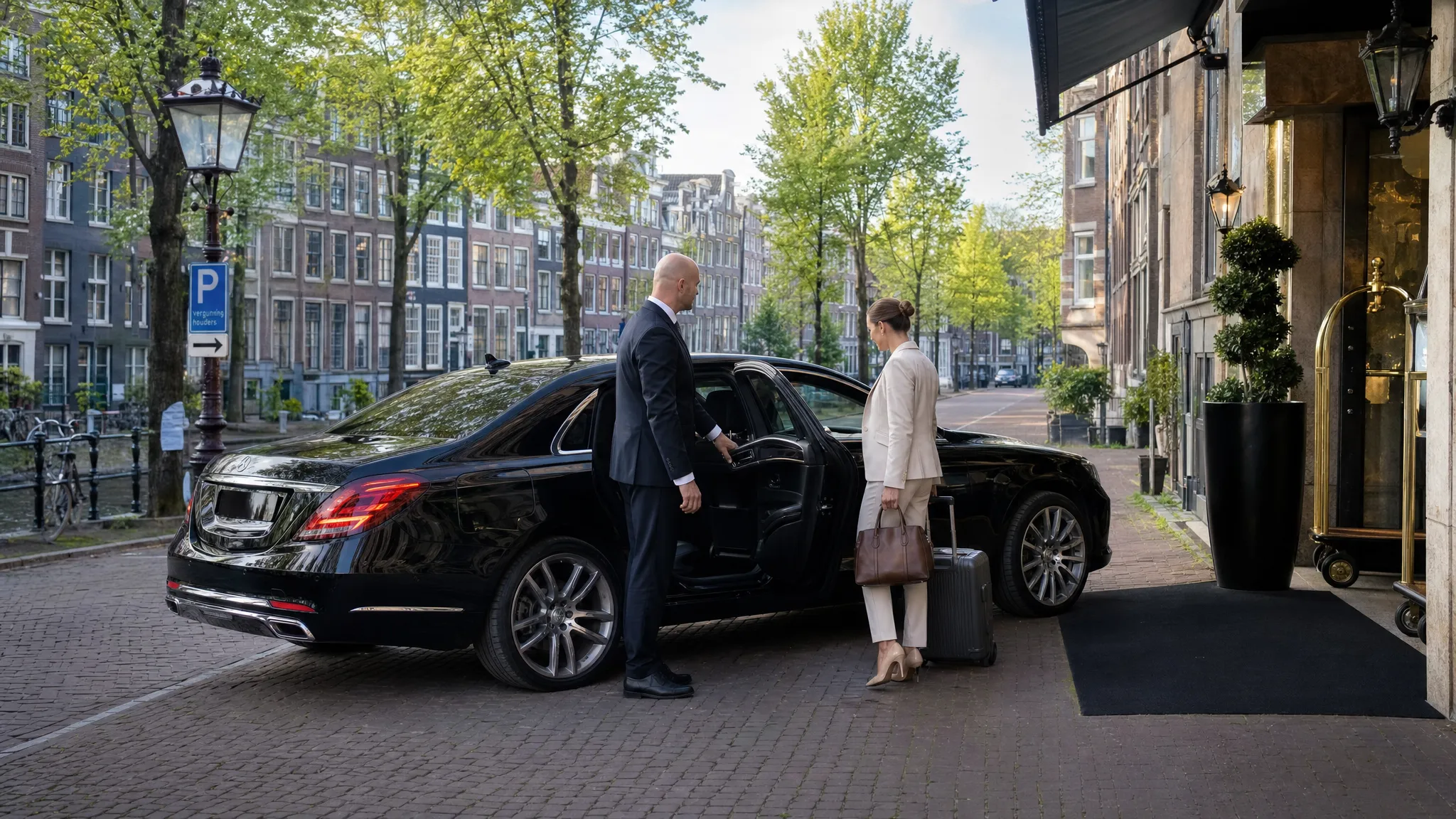 Een luxe zakelijke auto staat discreet bij een hotelentree in Amsterdam, met de chauffeur bij het achterportier en een passagier met handbagage naast de auto. Op de achtergrond zijn grachtenpanden en een rustige straat zichtbaar.