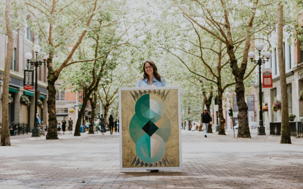 A woman stands in a city square holding a painting.