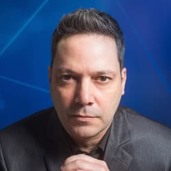 Professional portrait of a woman in a light tan blazer and purple blouse against a light gray wall.