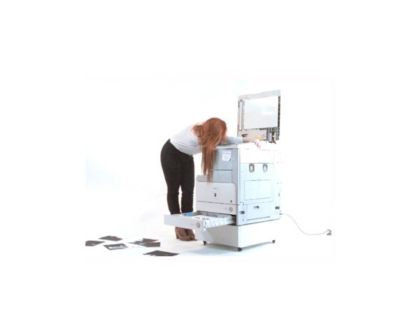 A woman with long reddish-brown hair, wearing a white long-sleeved shirt and black pants, bends over a large, light blue-gray machine against a white background. The machine has a pull-out drawer and a partially open lid revealing internal components. A black cable extends from the machine. The woman is positioned to the left of the machine, with her body angled slightly forward. Black rectangular objects are scattered on the floor near the machine’s base.