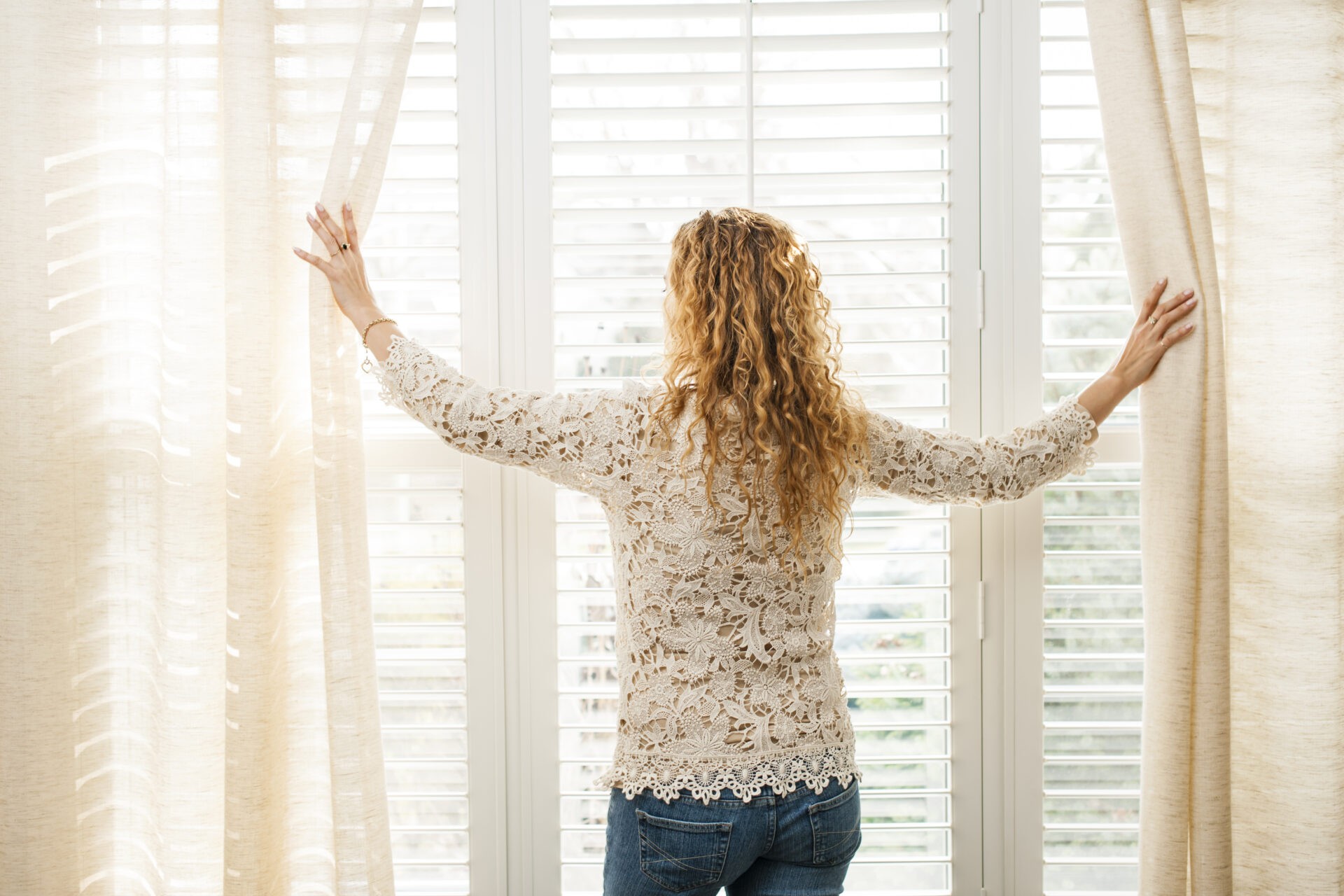 Woman looking out of shades with big bright window and Venetian shades. Wholesale Blind Factory.