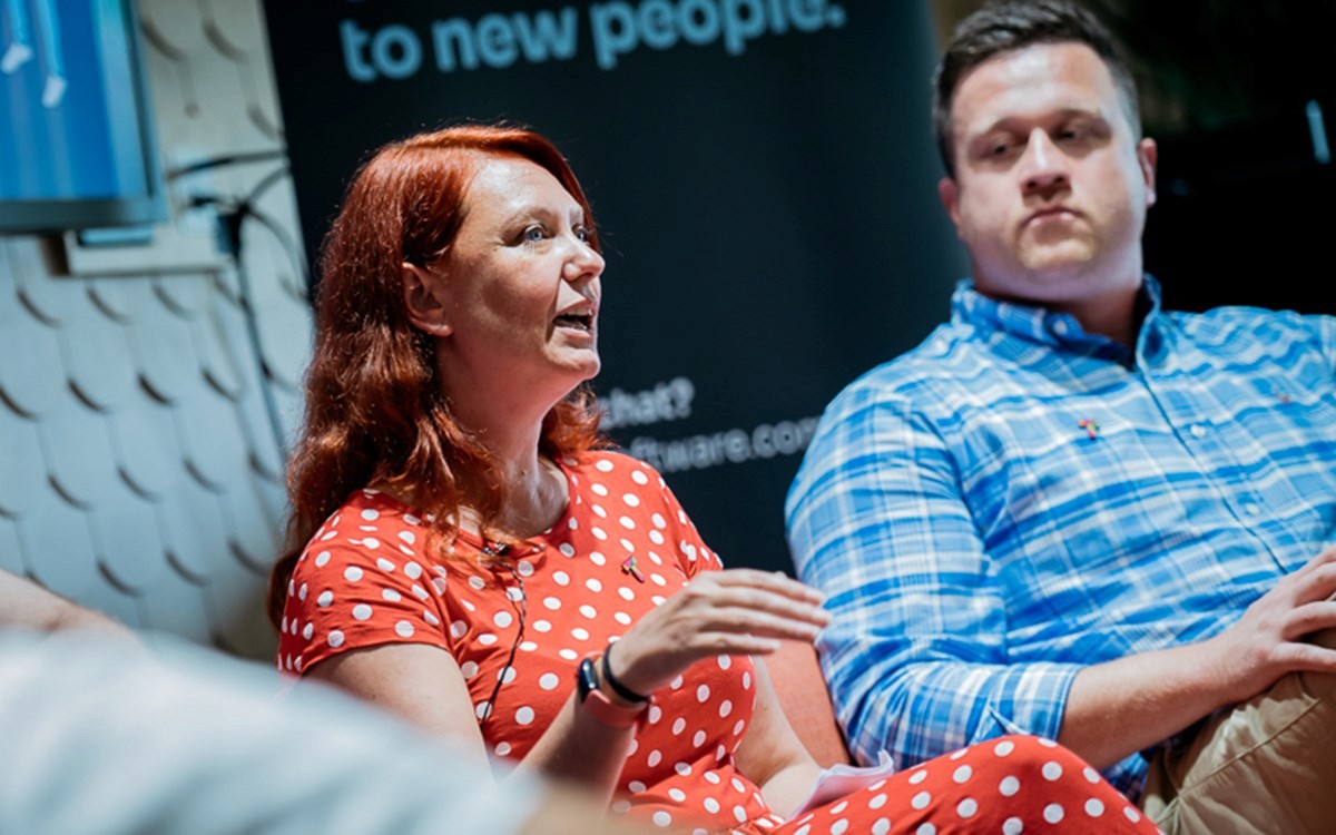 Two people seated indoors during a discussion, one wearing a red polka dot dress and the other in a blue plaid shirt.
