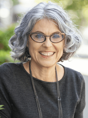 Woman wearing glasses and a floral blouse, sitting in natural light and smiling gently at the camera.