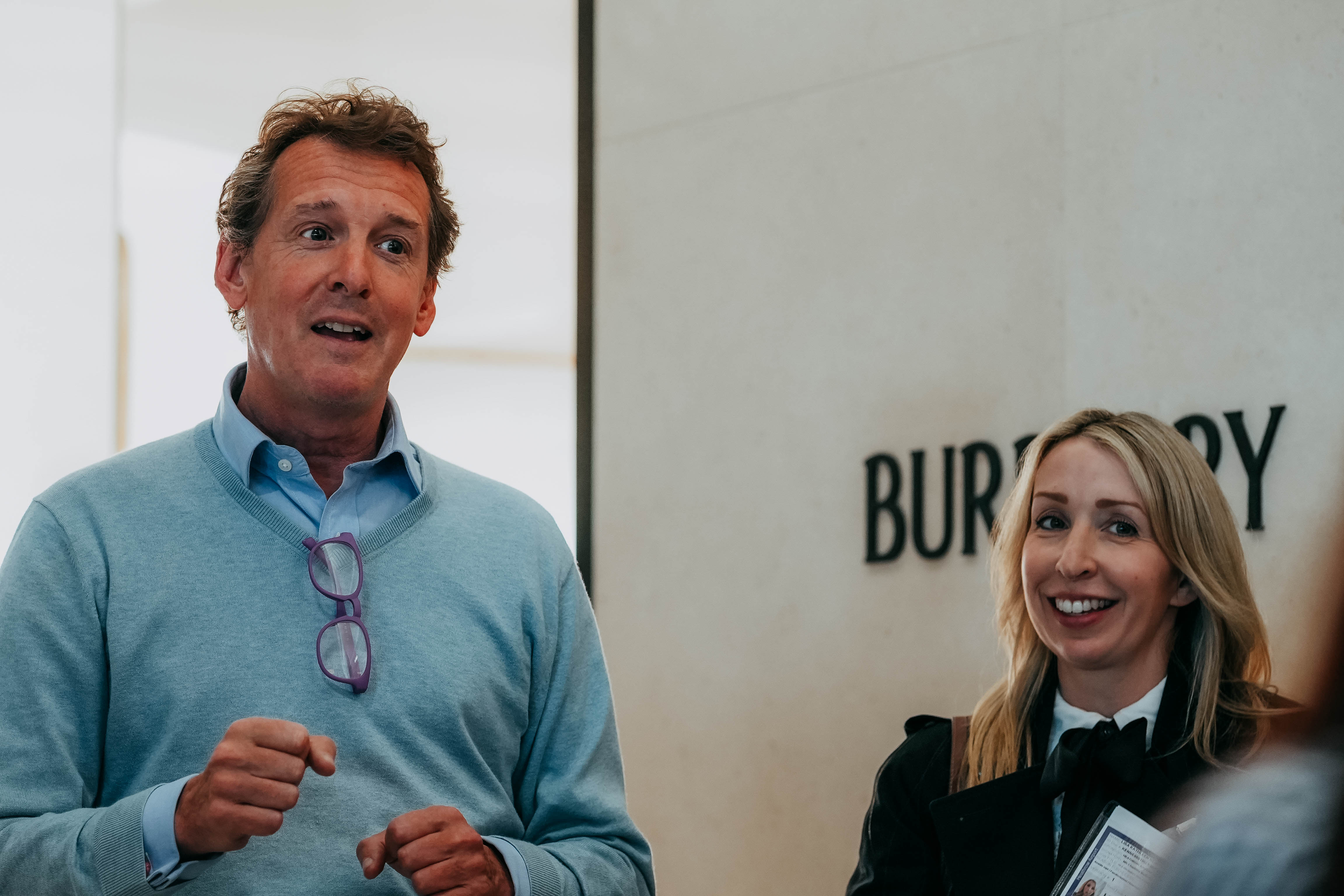 A man in a light blue sweater is speaking expressively while a blonde woman next to him smiles and listens. They are standing in front of a wall with the Burberry logo.