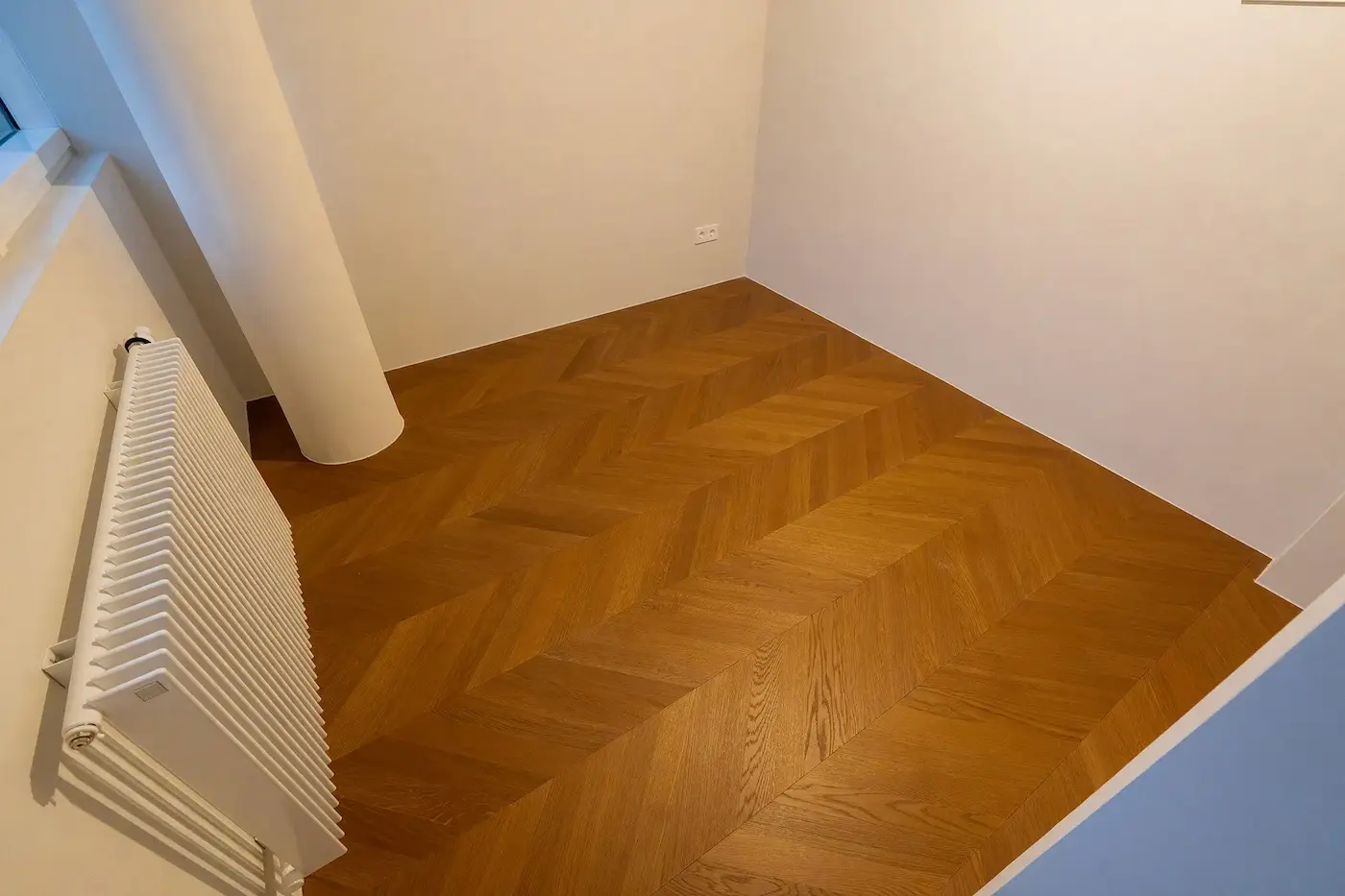 Detailansicht eines präzise verlegten Chevron-Parkettbodens entlang einer Wand und Säule