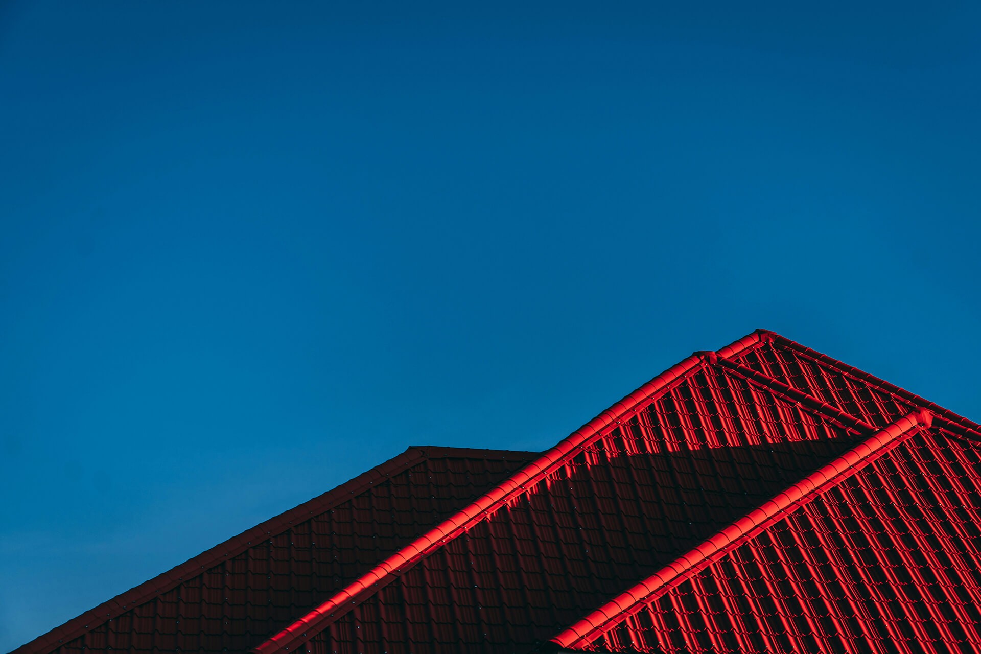 Close-up view of a red-tiled rooftop with sharp architectural angles, dramatically lit against a clear deep blue sky. The lighting emphasizes the texture and pattern of the roof tiles, suggesting a focus on roofing quality or design.