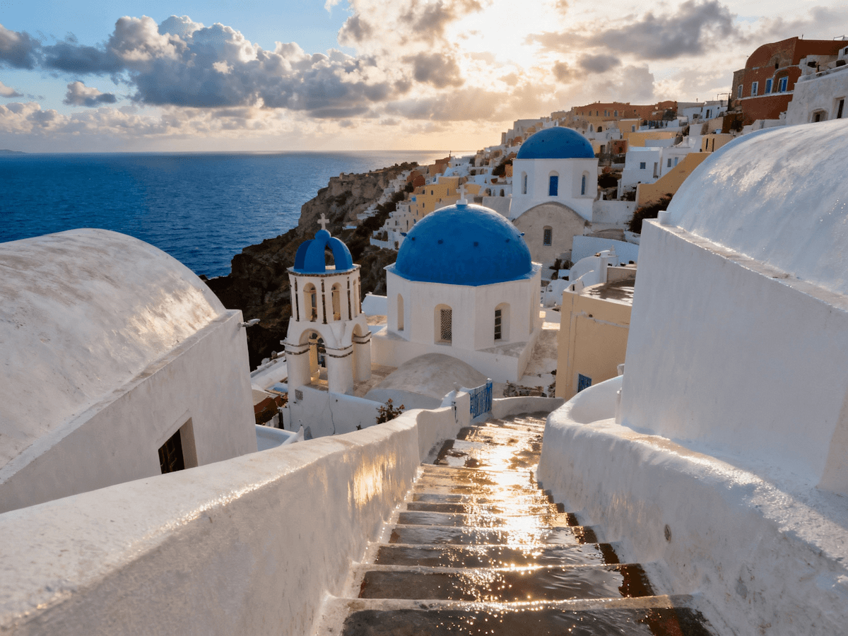 alt="Whitewashed buildings with blue domes overlooking the sea on a sunny coastal cliff."