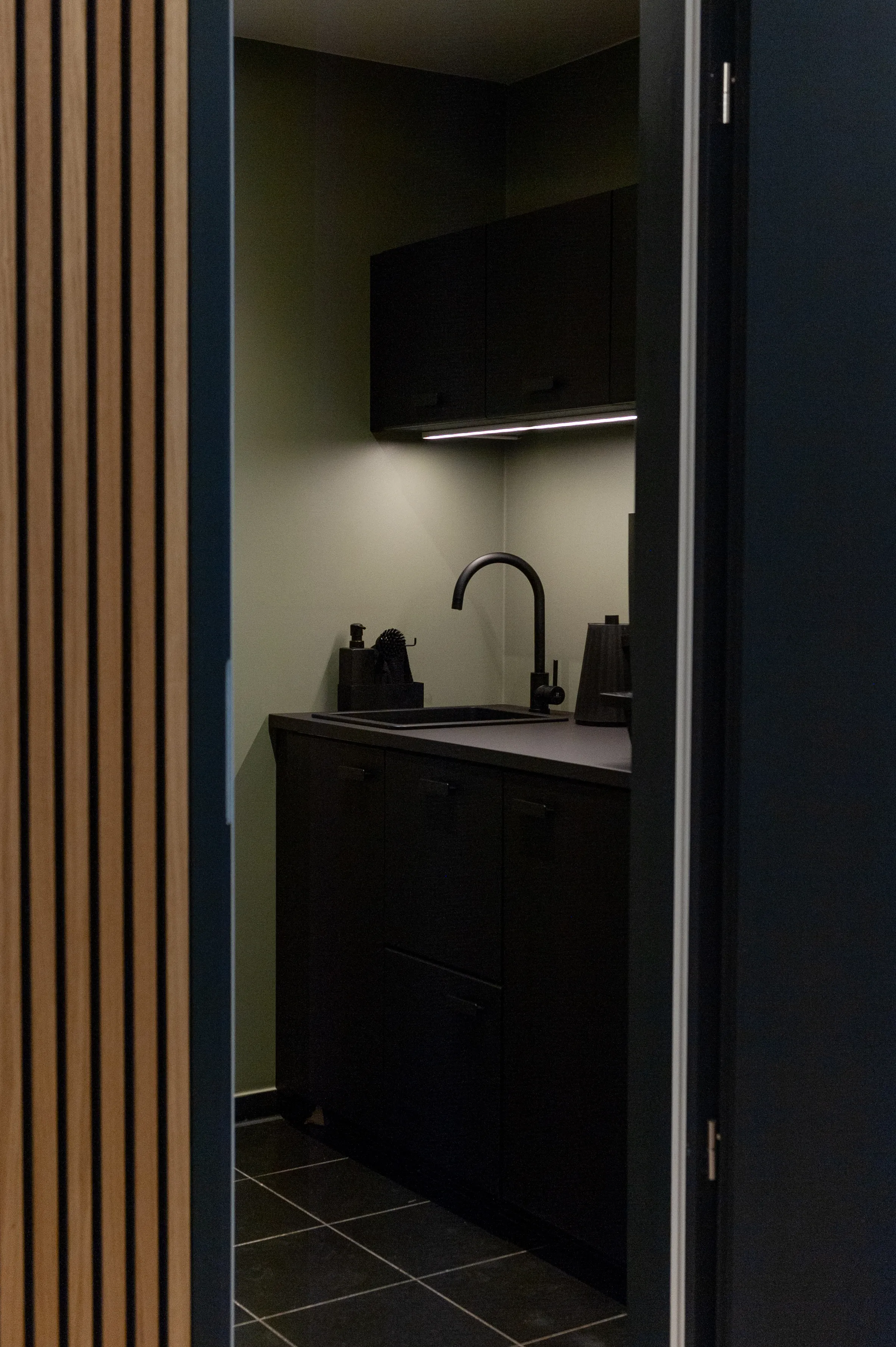 View through a doorway into the black kitchenette at AI START-UP BERLIN with sage-green walls, black cabinets, under-cabinet lighting and dark slate floor tiles, framed by an oak slat wall.