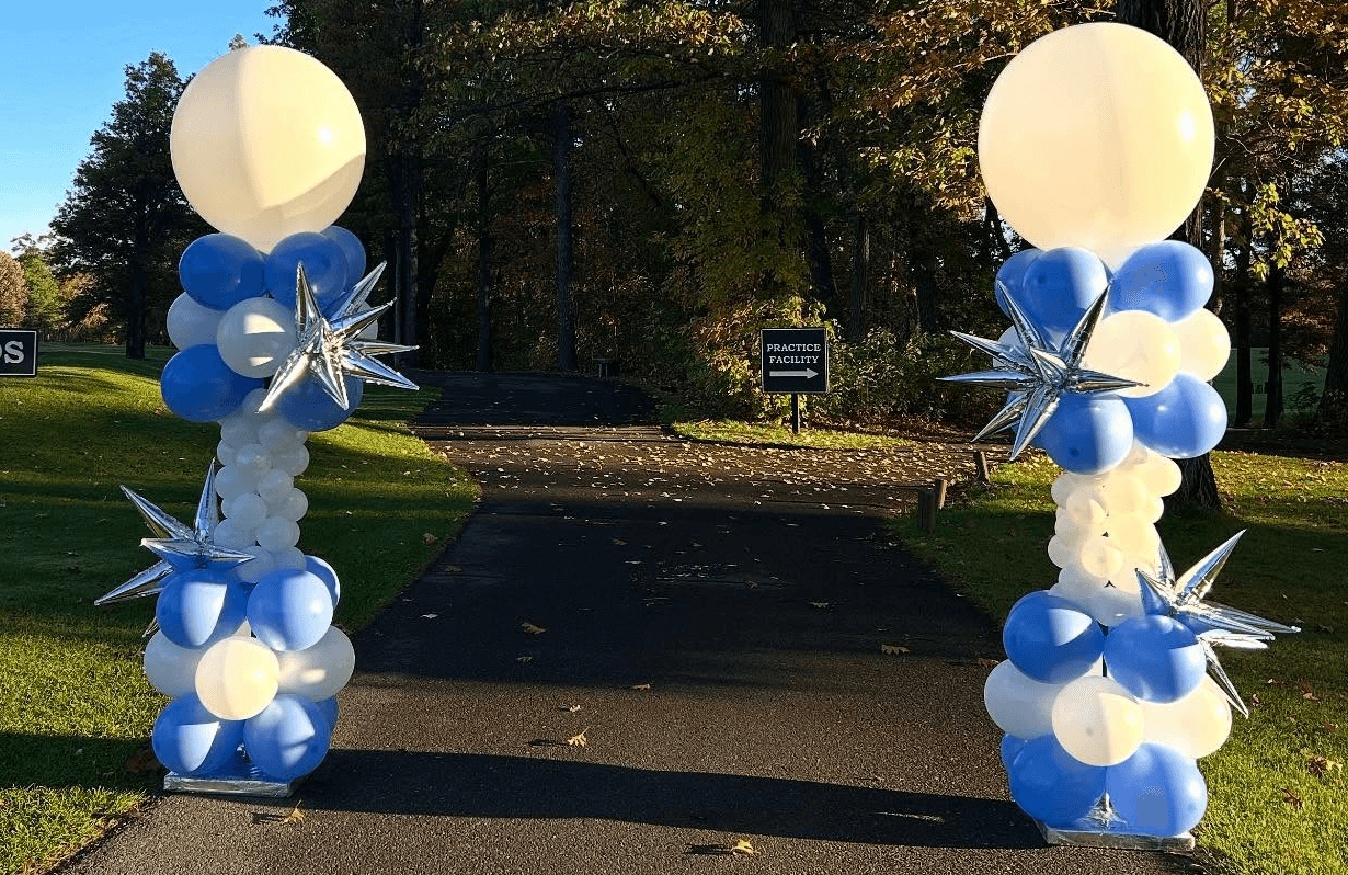 Blue and white balloon columns with large white balloon toppers framing a park entrance or walkway by Be Bold Balloons