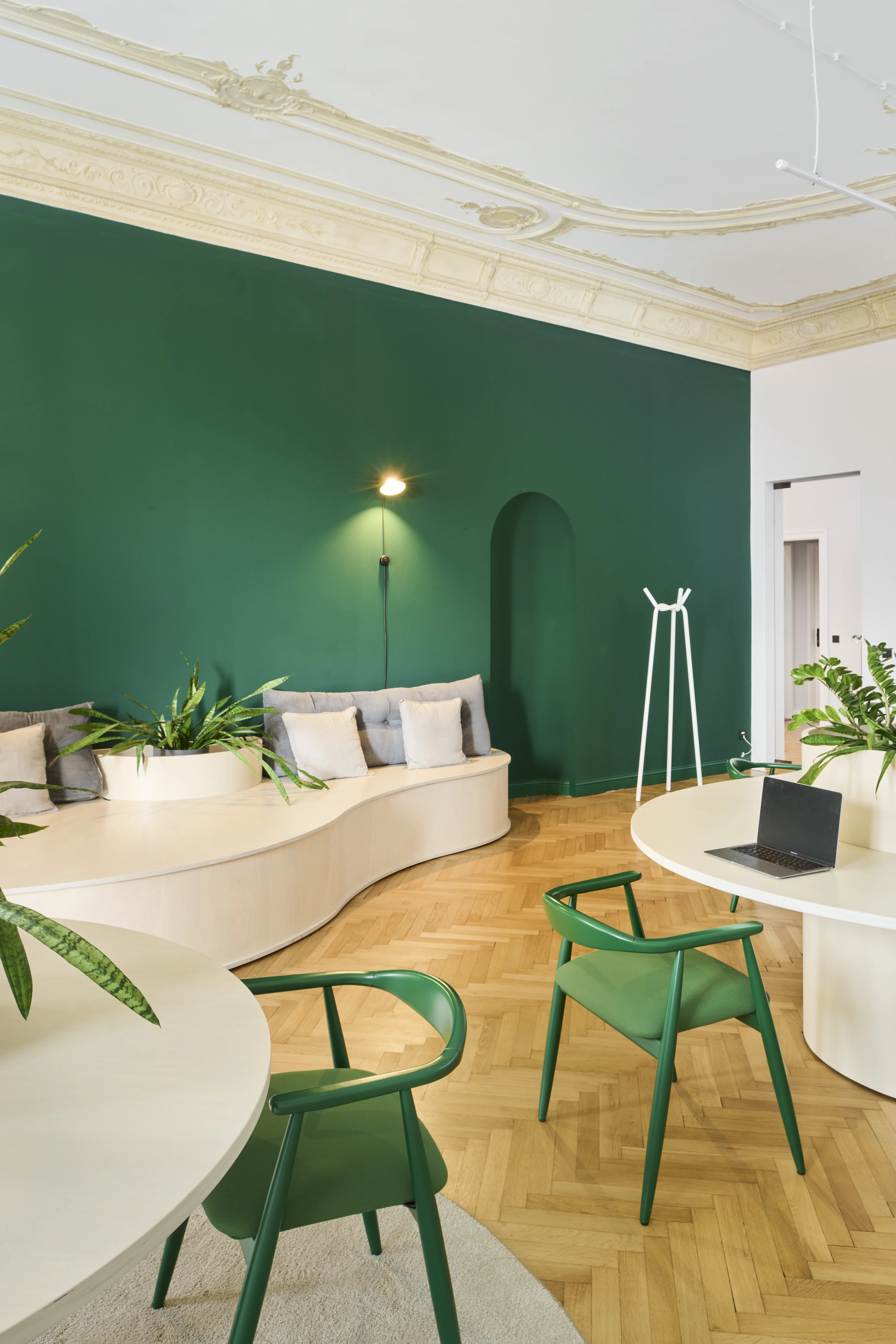 Green salon at INTERMATE BERLIN with an emerald wall with arched niche, a curved built-in bench with cushions and planters, green wooden chairs, white round tables and stucco ceiling.