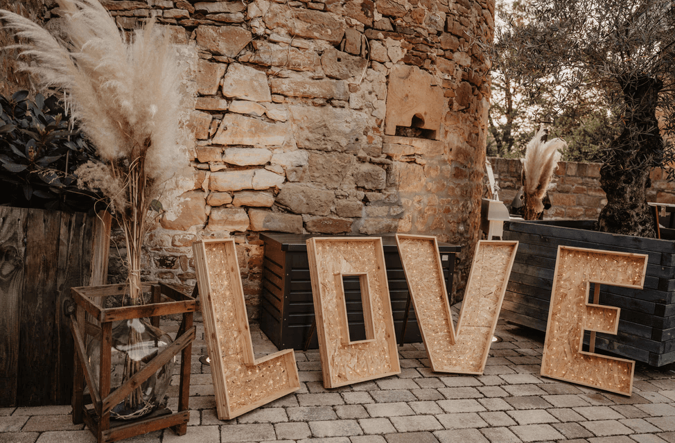 XXL Leuchtbuchstaben 'LOVE' aus Holz mit Glühbirnen vor einer rustikalen Steinfassade, dekoriert mit Pampasgras.