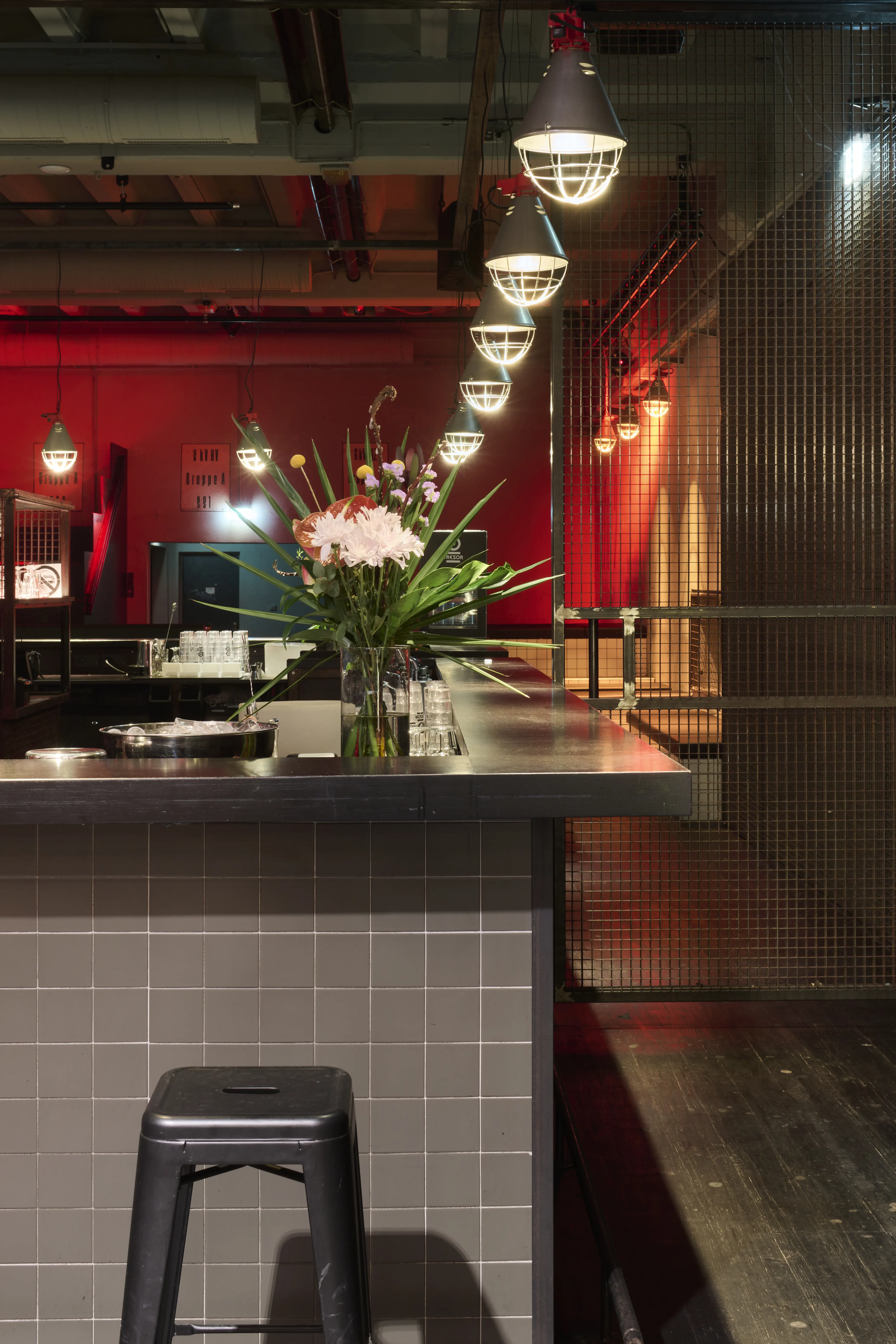 Bar counter at TRESOR GLOBUS with grey tiled base, black top, fresh flowers and a row of caged industrial pendant lights against a red-lit room.