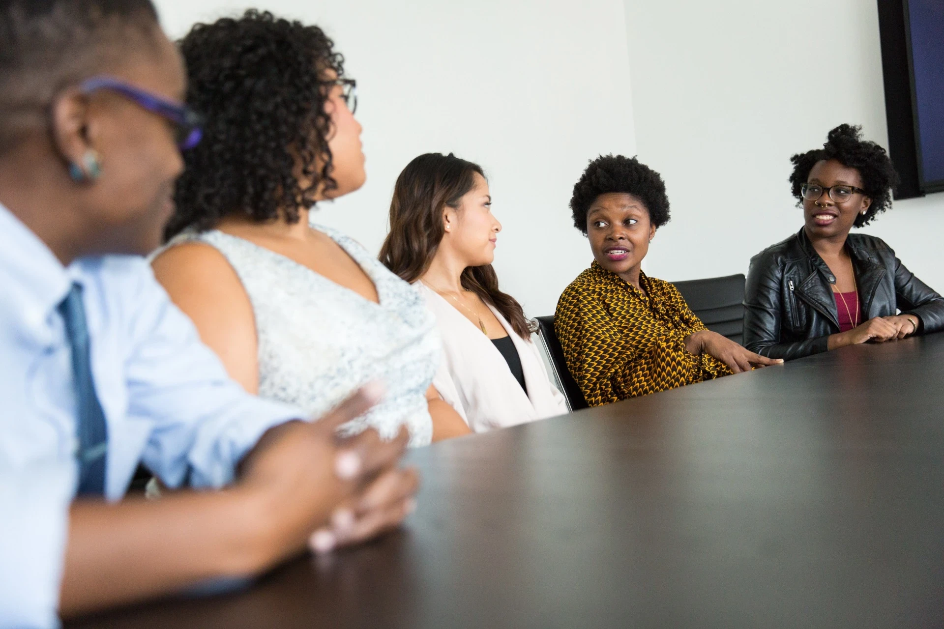 Imagem de um grupo diversificado de pessoas sentadas ao redor de uma mesa de reunião, engajadas em uma discussão. Uma mulher de blusa estampada parece estar falando, enquanto os outros a escutam atentamente, cada um com expressões diferentes que refletem interesse e atenção. A cena representa um ambiente de colaboração e troca de ideias, enfatizando a diversidade e a interação profissional em um ambiente de trabalho inclusivo.