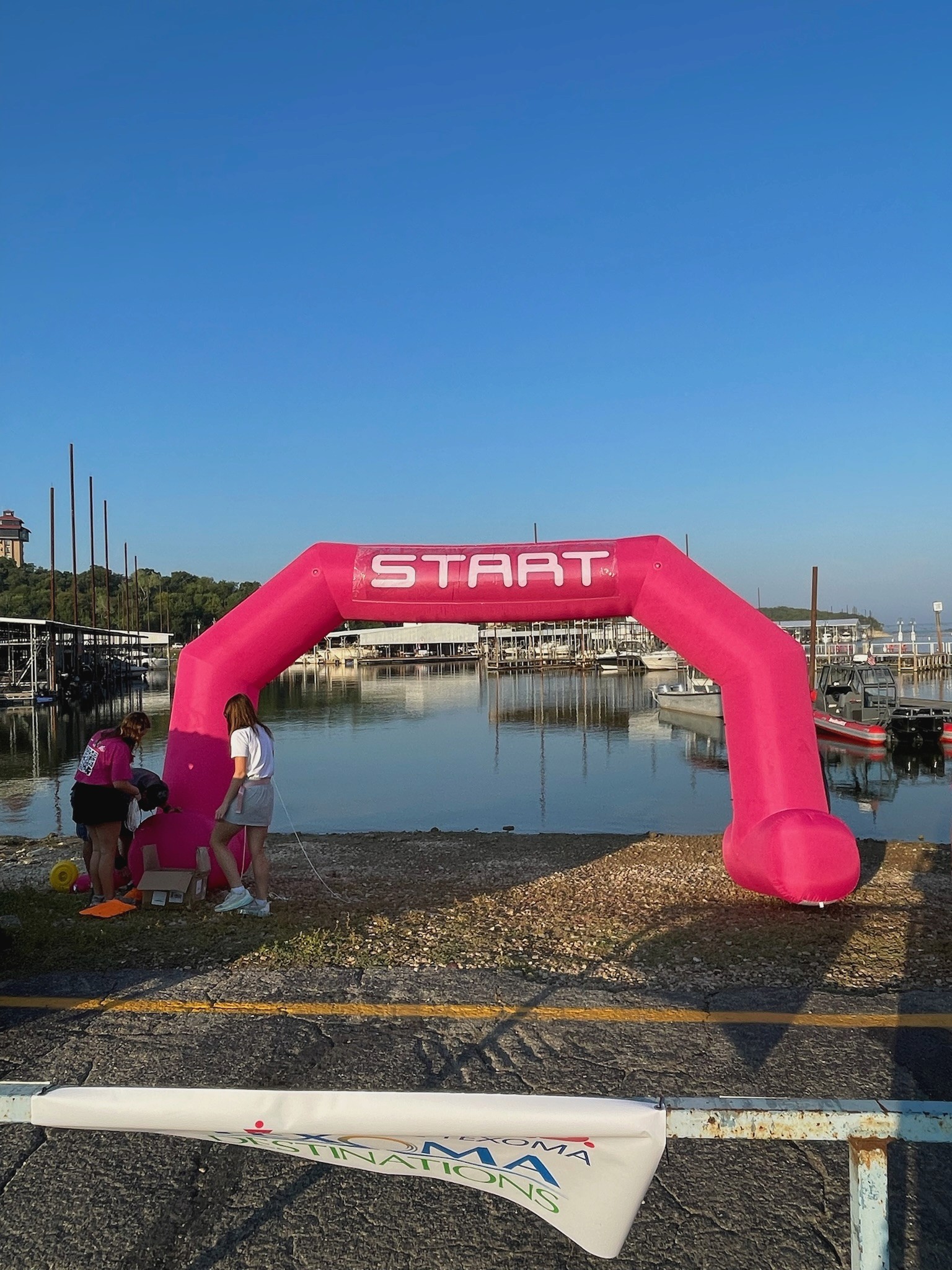 A vibrant pink inflatable arch labeled "START" is set up by the water's edge at a marina, with boats docked in the background on a clear, sunny day.