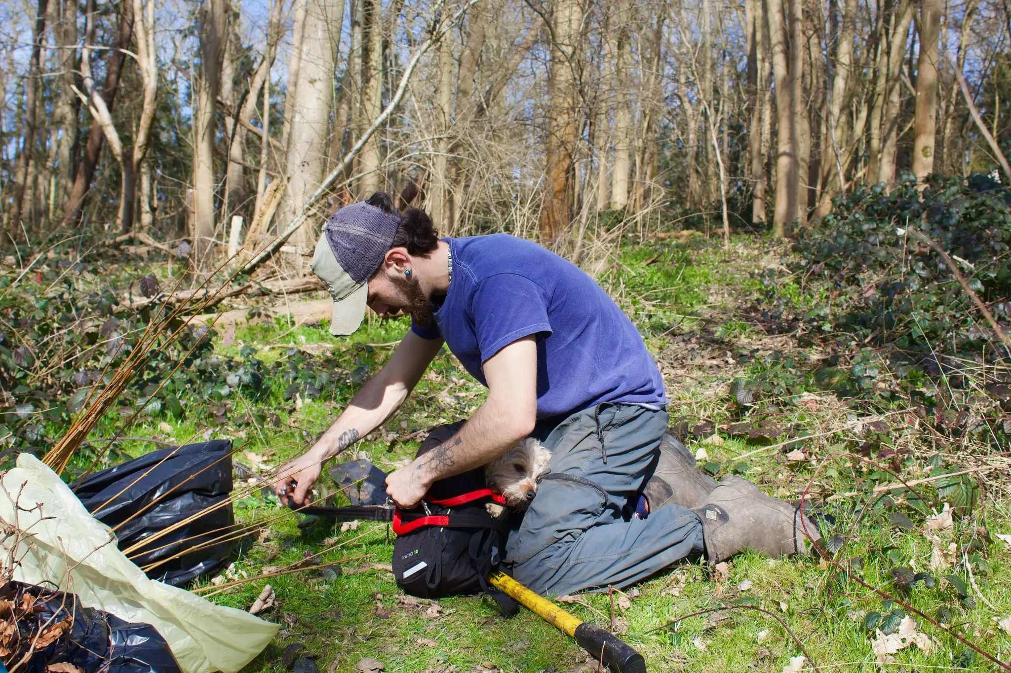 A person kneels on the ground in a forest, studying materials spread on the grass.