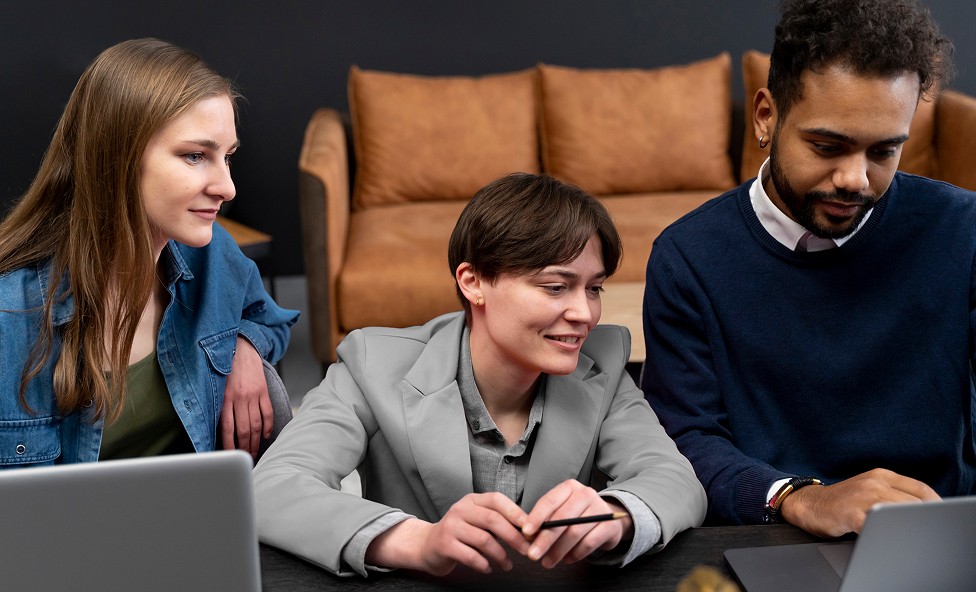 Three people sit at a table, collaborating while intently looking at laptops, with a brown sofa in the background, conveying a professional office setting.