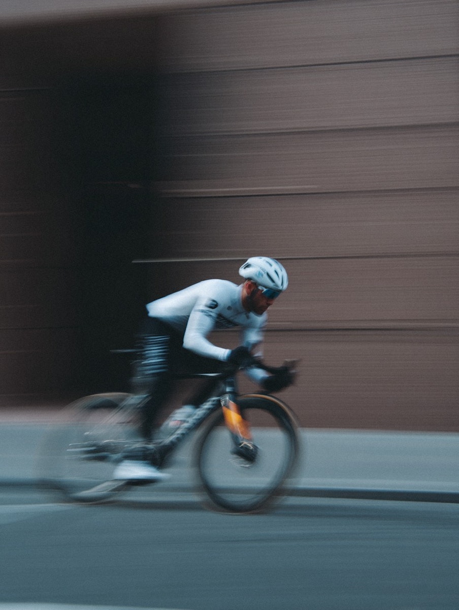 A cyclist in motion, wearing a white jersey and helmet, rides a sleek black bike with orange accents against a blurred urban backdrop.