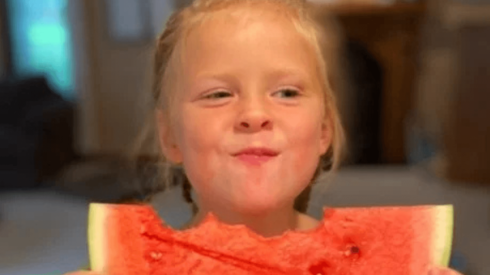 Young child smiling while eating a slice of watermelon indoors.