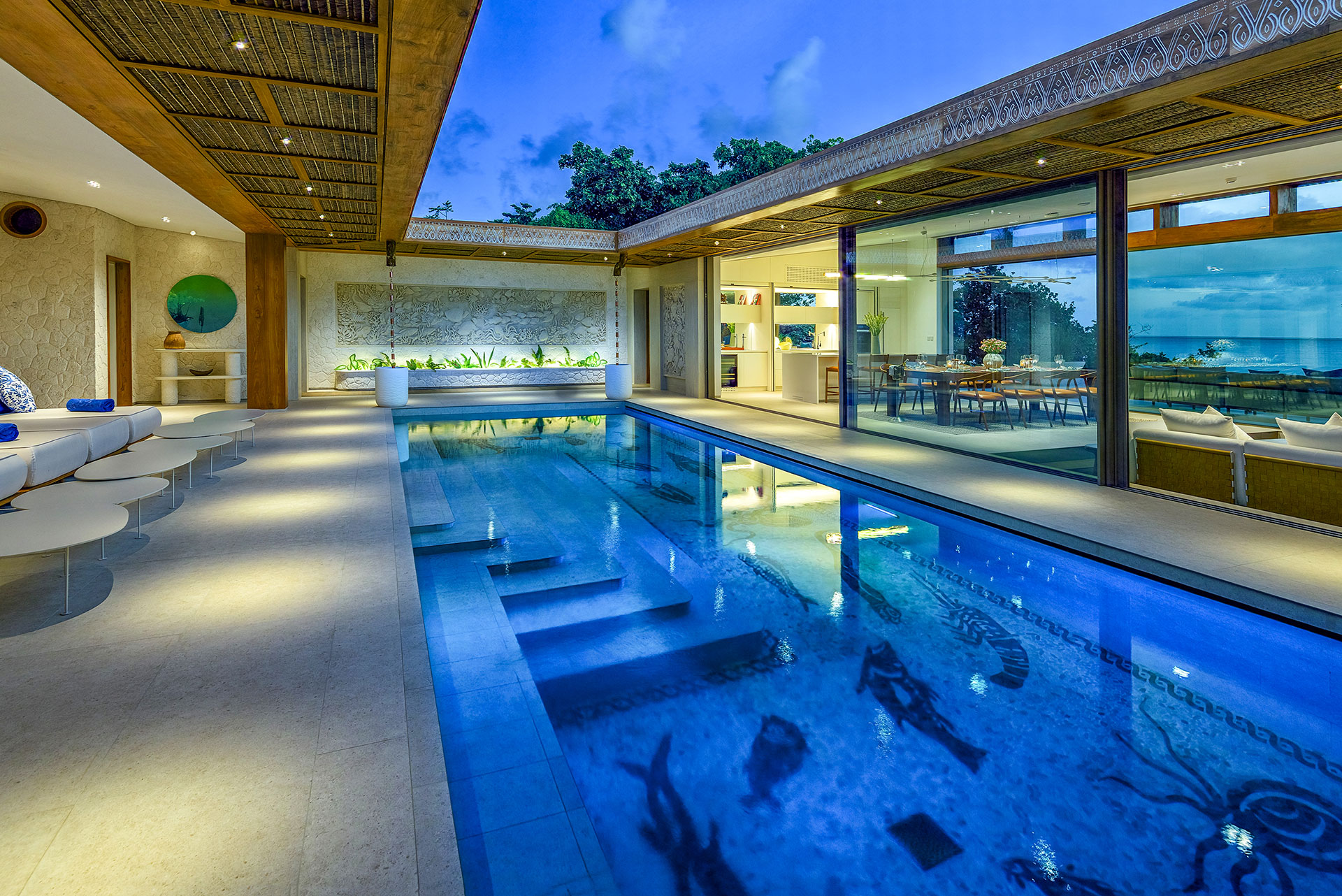 Modernes Atrium bei Dämmerung mit einem zentralen Pool, dessen Boden mit dunklen Mosaiken von Meerestieren (wie Hummern und Fischen) verziert ist, flankiert von beleuchteten Wohnbereichen mit Glaswänden.