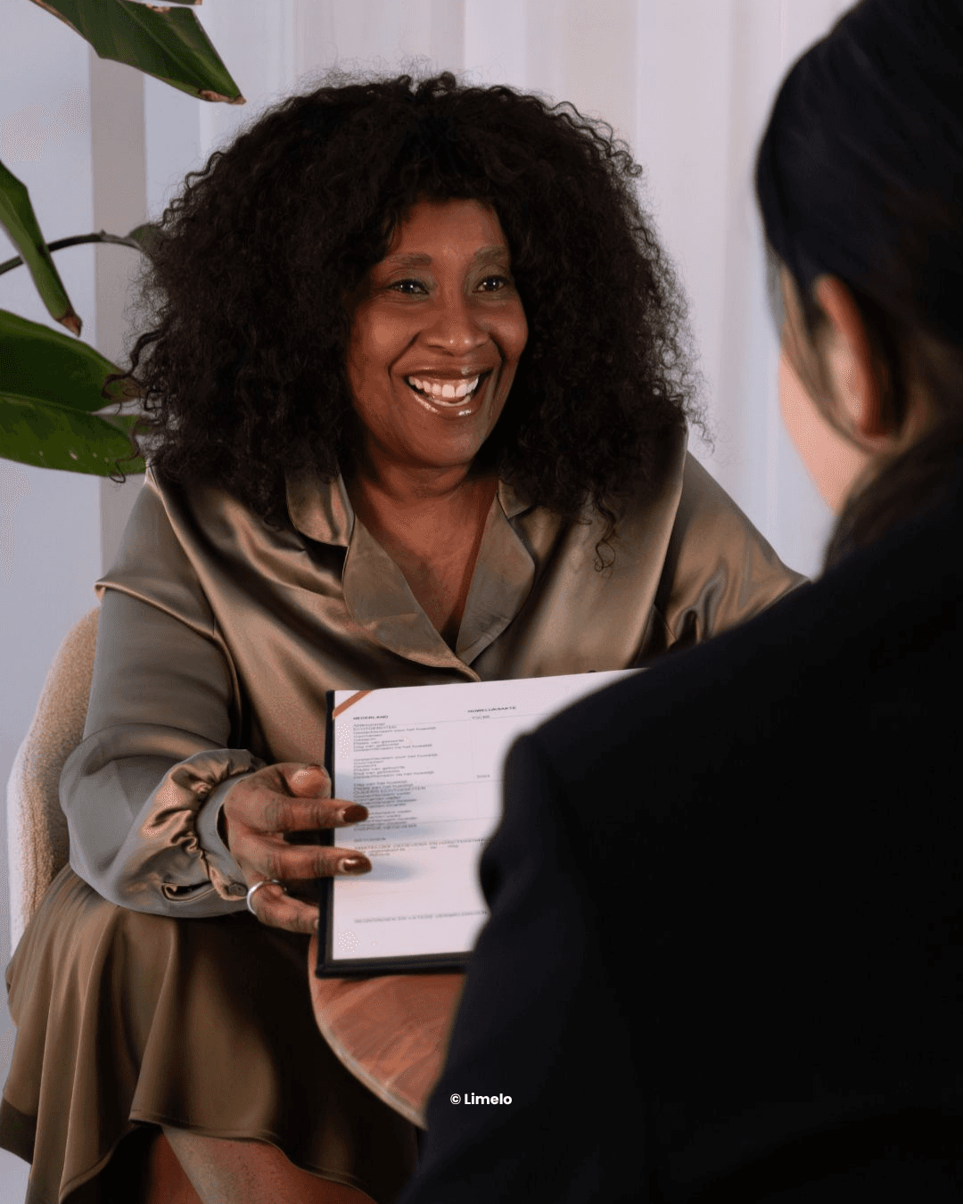 Een trouwambtenaar met krullend haar glimlacht terwijl ze een document aan een ander overhandigt en in gesprek is.