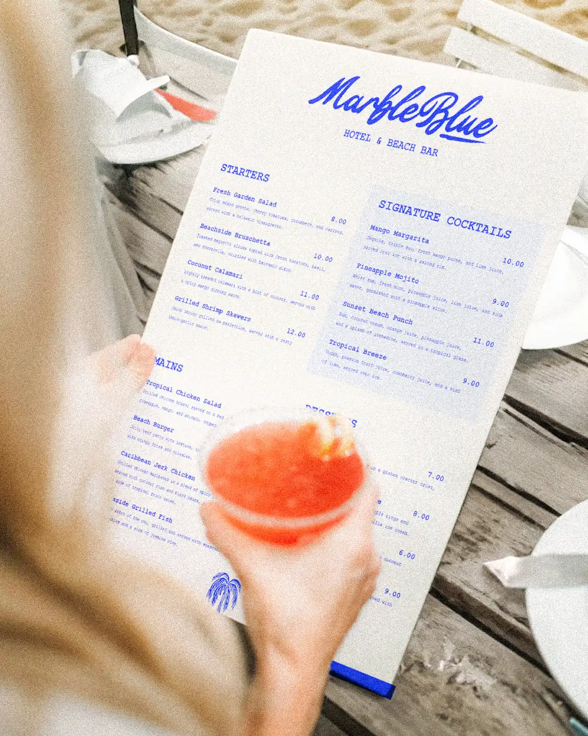 Menu Marble Blue tenu à la main sur la plage, avec cocktail rouge au premier plan flou, cartes starters, mains et signature cocktails visibles.