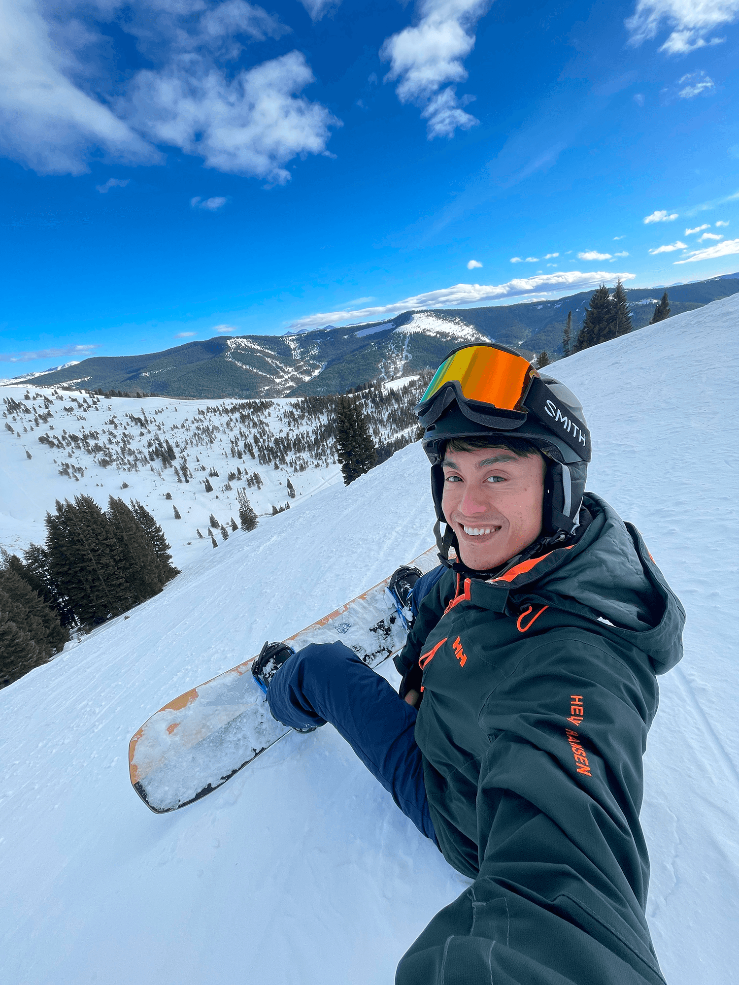 Snowboarder on top of a snow covered mountain with mountain ranges and tress in the background.