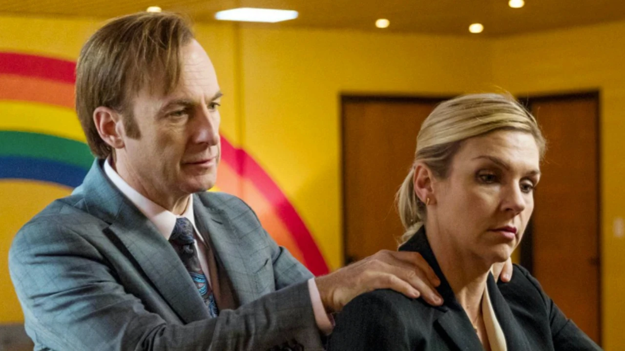 Dans un bureau aux murs jaunes ornés d’un motif arc-en-ciel, un homme en costume gris se tient derrière une femme en veste noire et pose ses mains sur ses épaules. La femme regarde vers le bas, le visage fermé, traduisant une tension ou un malaise. Cette scène de la série TV “Better Call Saul” met en lumière les rapports de pouvoir, la communication implicite et les dynamiques internes, en écho à la réflexion sur la culture d’entreprise comme socle invisible de la marque.