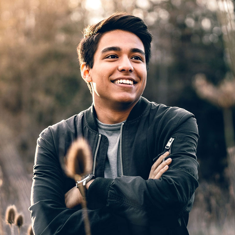 Fotografía de un hombre joven de piel morena y cabello negro, con vestimenta casual de chaqueta de cuero. El fondo desenfocado de árboles resalta al sujeto, representando el perfil de un abogado joven en un entorno natural y relajado.