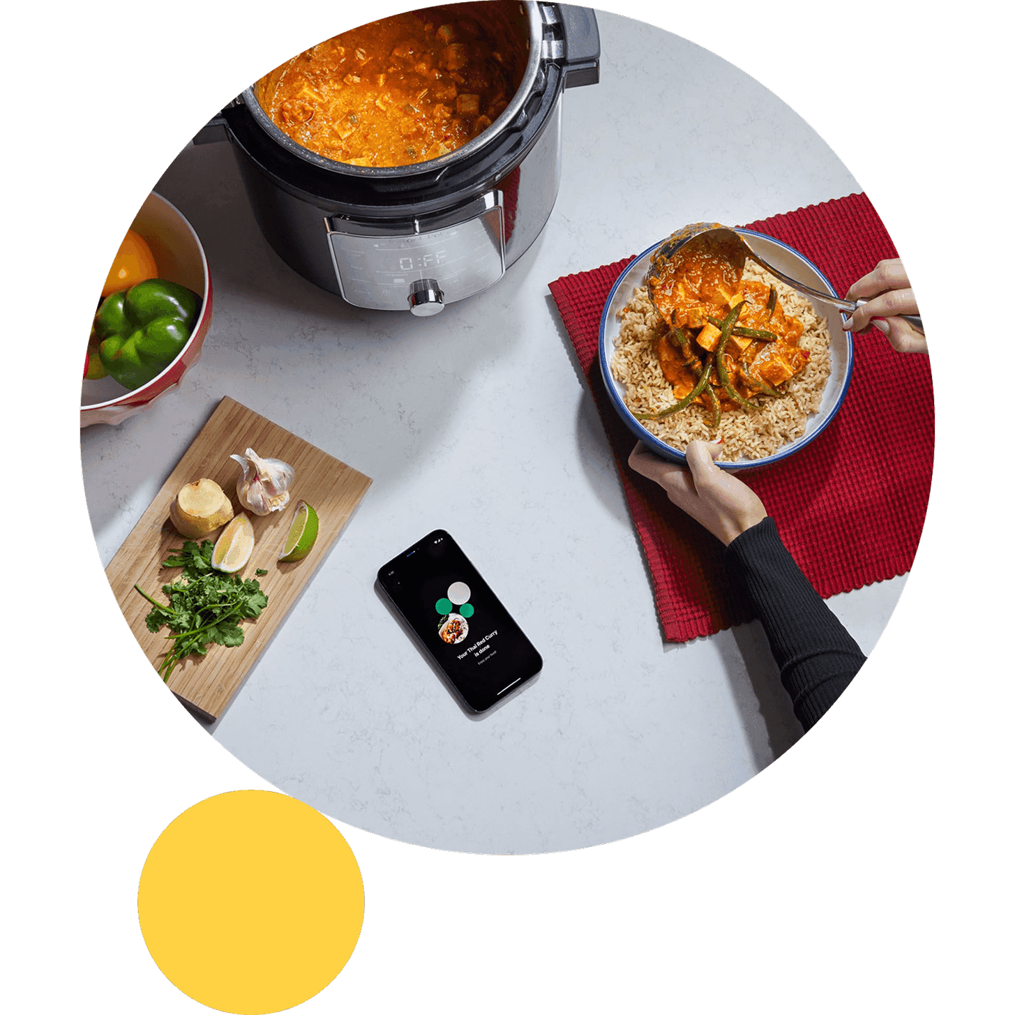 Top down view of a multicooker filled with curry beside a bowl of rice being served, with fresh ingredients on a cutting board and a smartphone app nearby