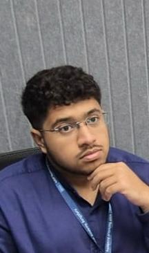 A person with curly hair and glasses smiles at the camera, seated in a cozy workspace with warm lighting.