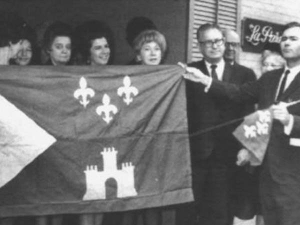 A group of people are gathered around a large dark flag with a white fleur-de-lis and a white castle emblem. The flag is held aloft by two individuals, one on each side. The group includes several women and at least one man. The man on the right is wearing a dark suit and tie and is pointing toward the flag. The background shows a building with horizontal siding and a sign reading “La Ferme”. The image is in black and white.