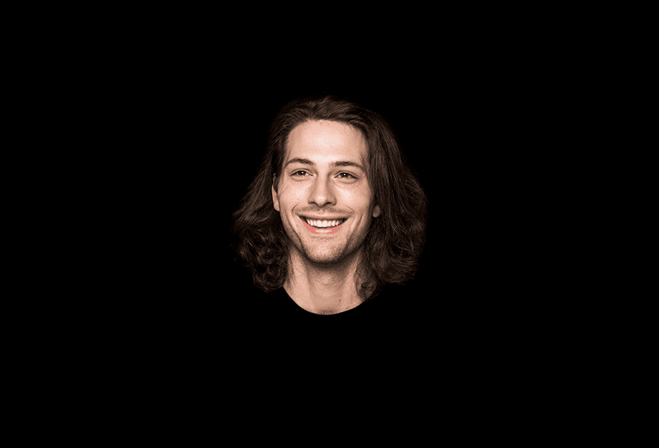 A man with long brown hair and a light complexion is smiling at the camera against a solid black background. He is wearing a dark-colored shirt.