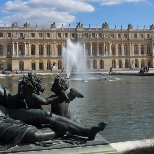 Statues of cherubs by a large fountain in front of the Palace of Versailles, with a clear sky above.