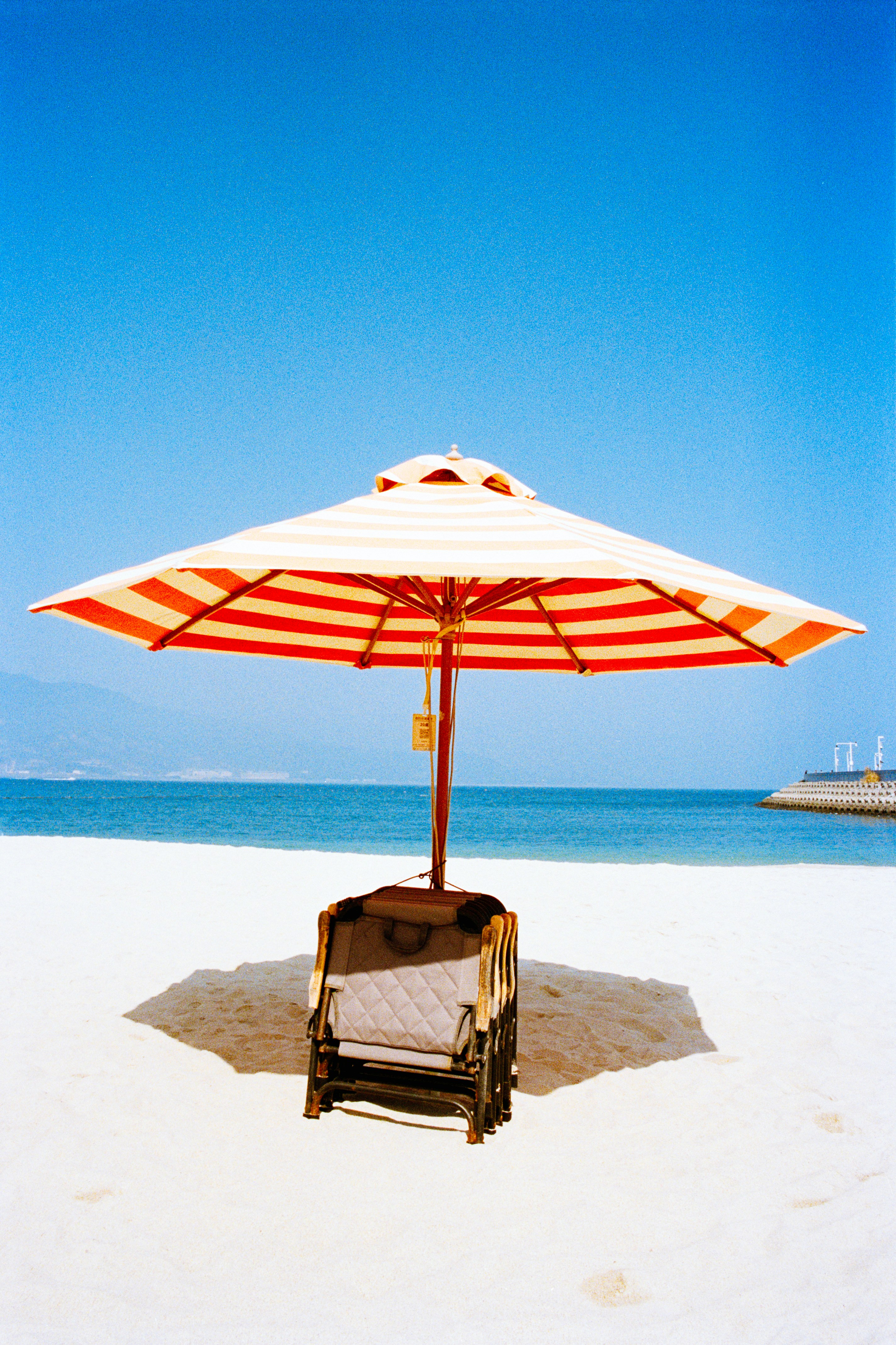 Striped beach umbrella with lounge chair on sand.