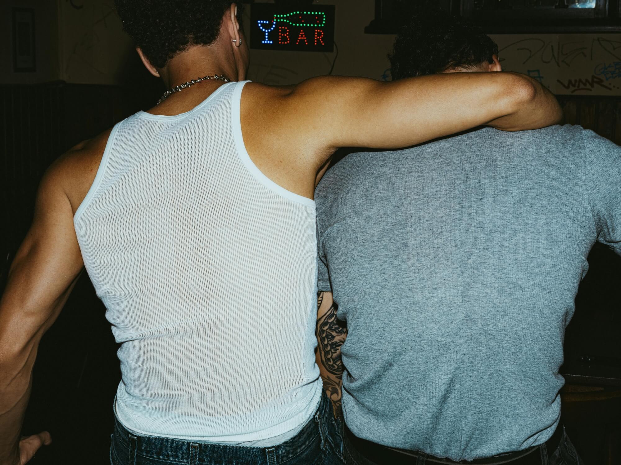 Two people with their backs turned, one in a white tank top and the other in a gray shirt, embrace in a dim bar with a neon "BAR" sign.