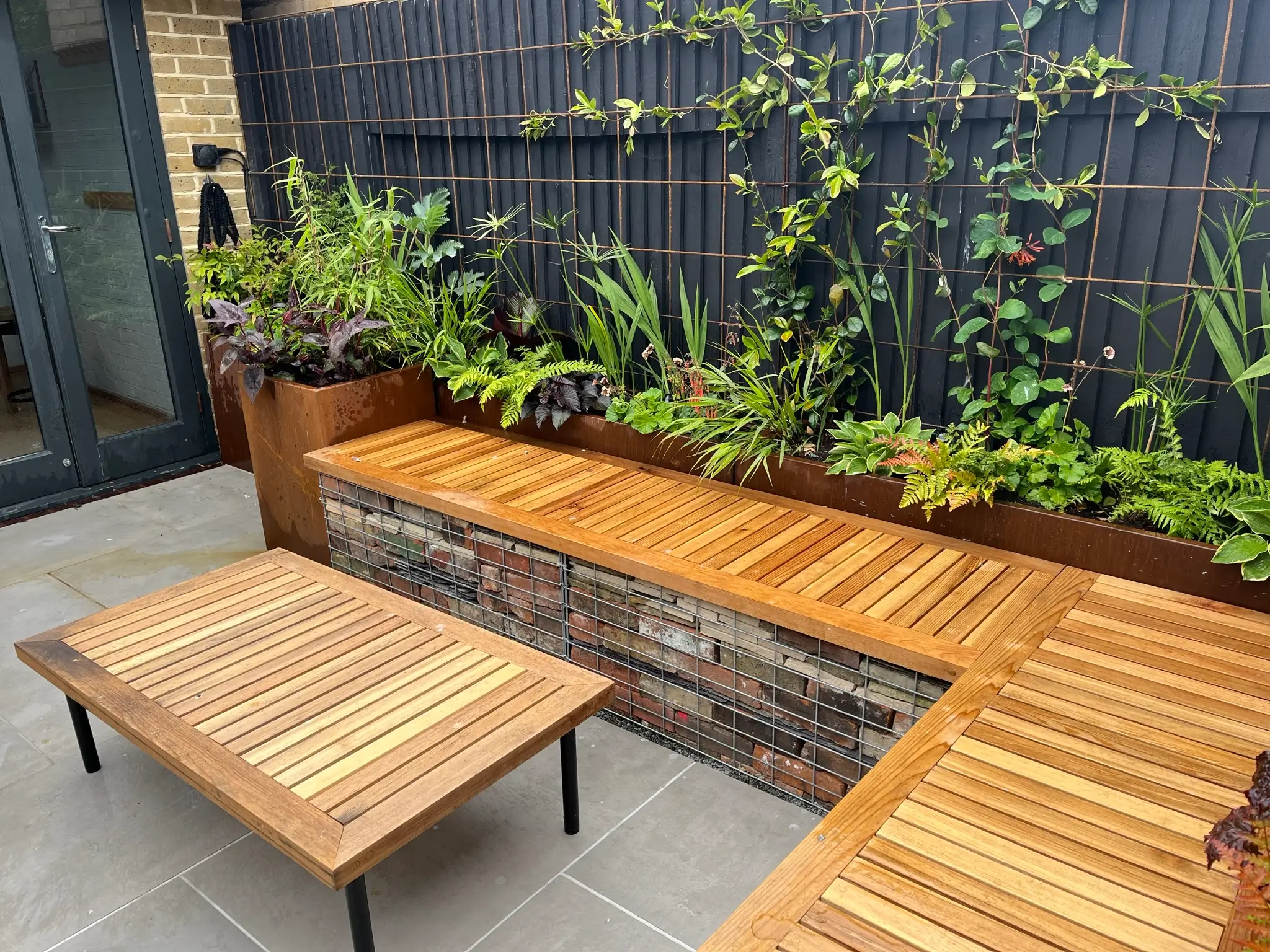 A cozy outdoor space with wooden seating, a low table, and lush greenery in planters along the wall.