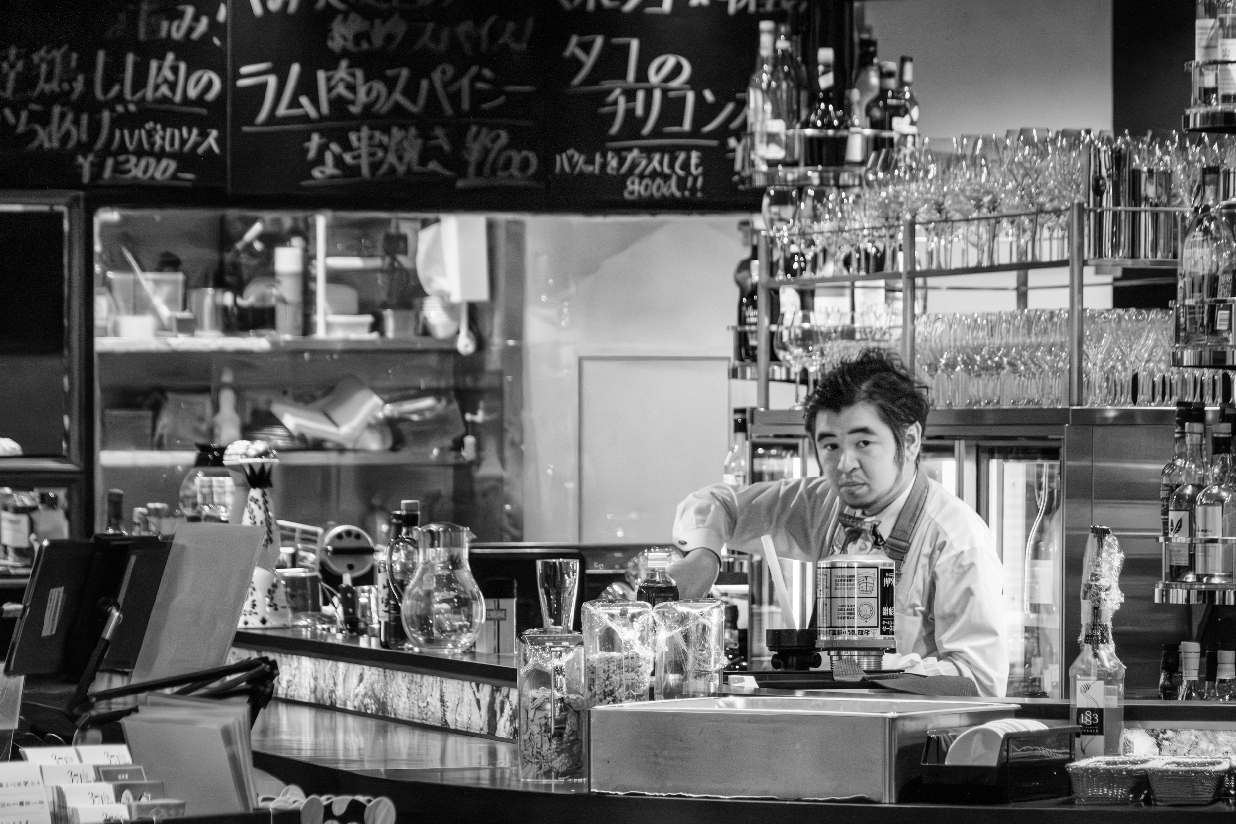 A black-and-white image of a person working at a counter surrounded by shelves filled with various items.