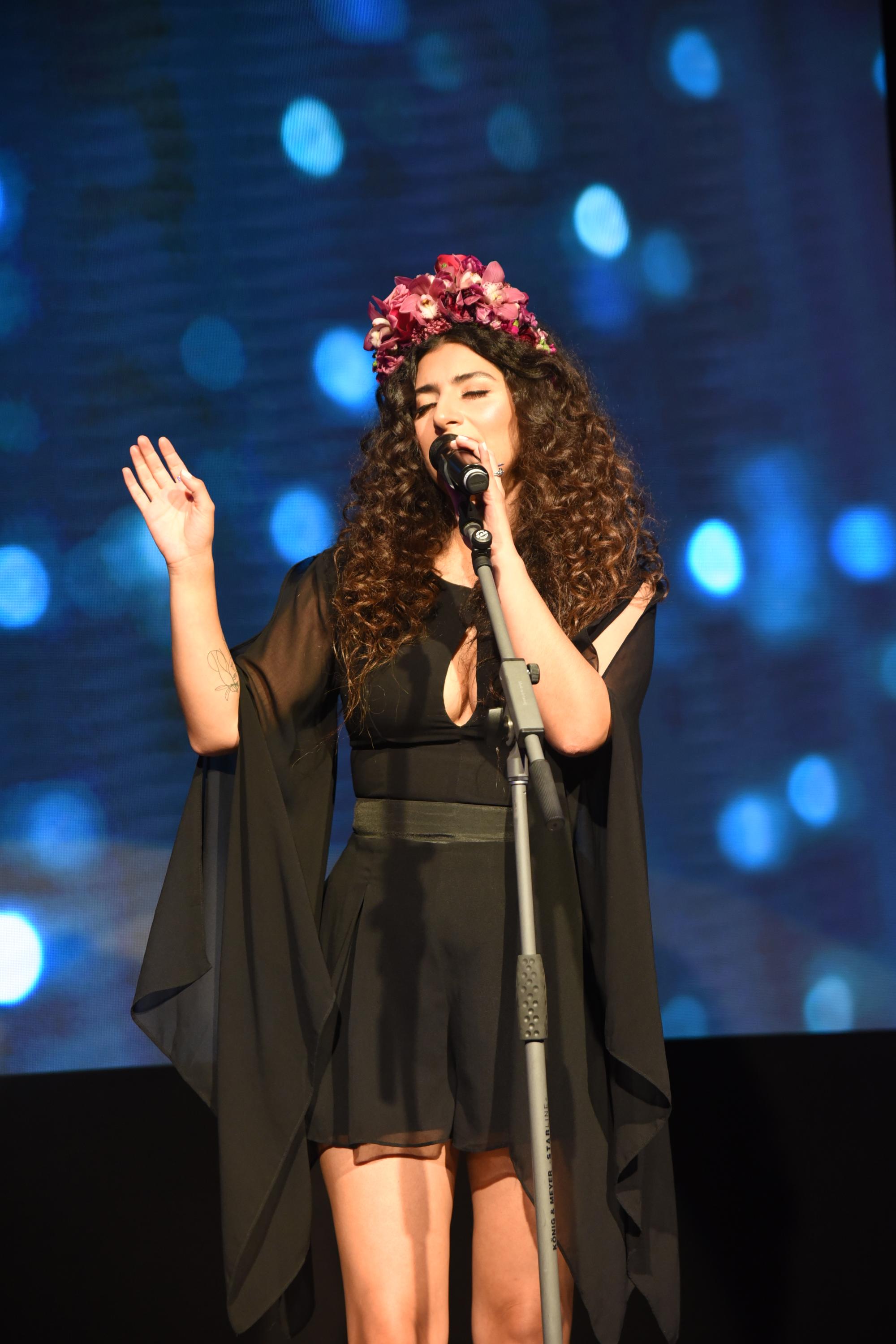Singer performing on stage with microphone and floral headpiece against a blue lit backdrop.
