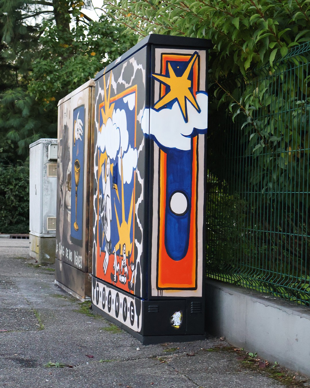 A painted electrical box with a sun above white clouds over an abstract orange-and-blue pattern on the side, with another painted electrical box partially visible behind it.