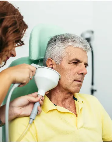 A healthcare professional uses a device on an older man's ear in a clinical setting.