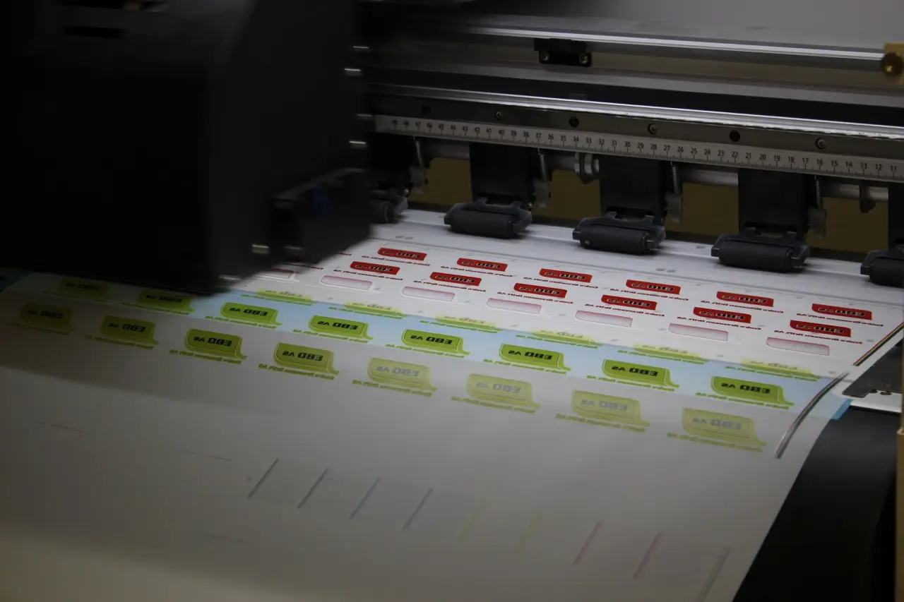 En stor trykkmaskin skriver ut et ark fylt med fargerike, rektangulære etiketter i rader, hver med tekst, som viser presisjon og industriell produksjon.
