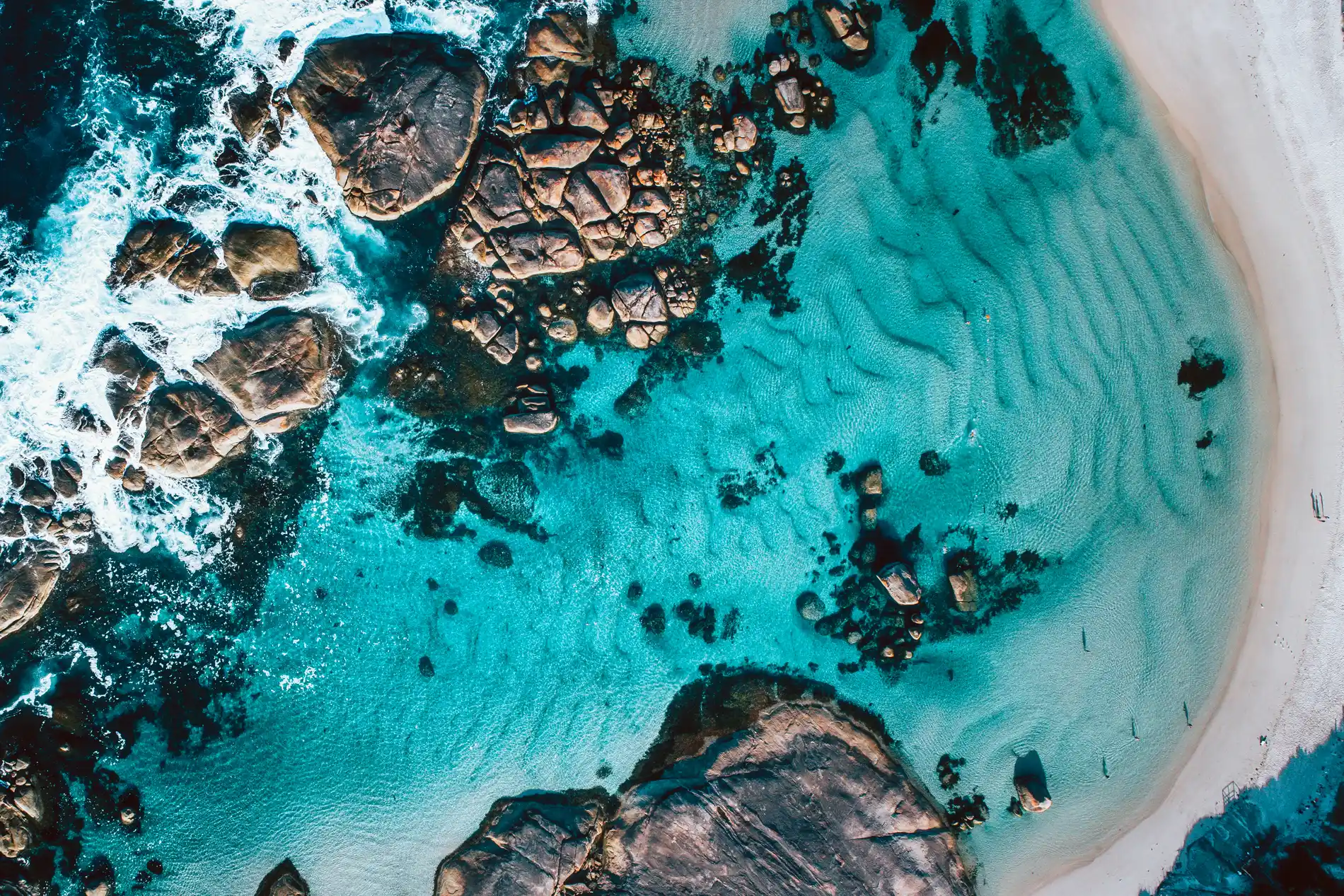 Aerial of dark reef and granite outcrops in bright turquoise shallows beside a curving beach