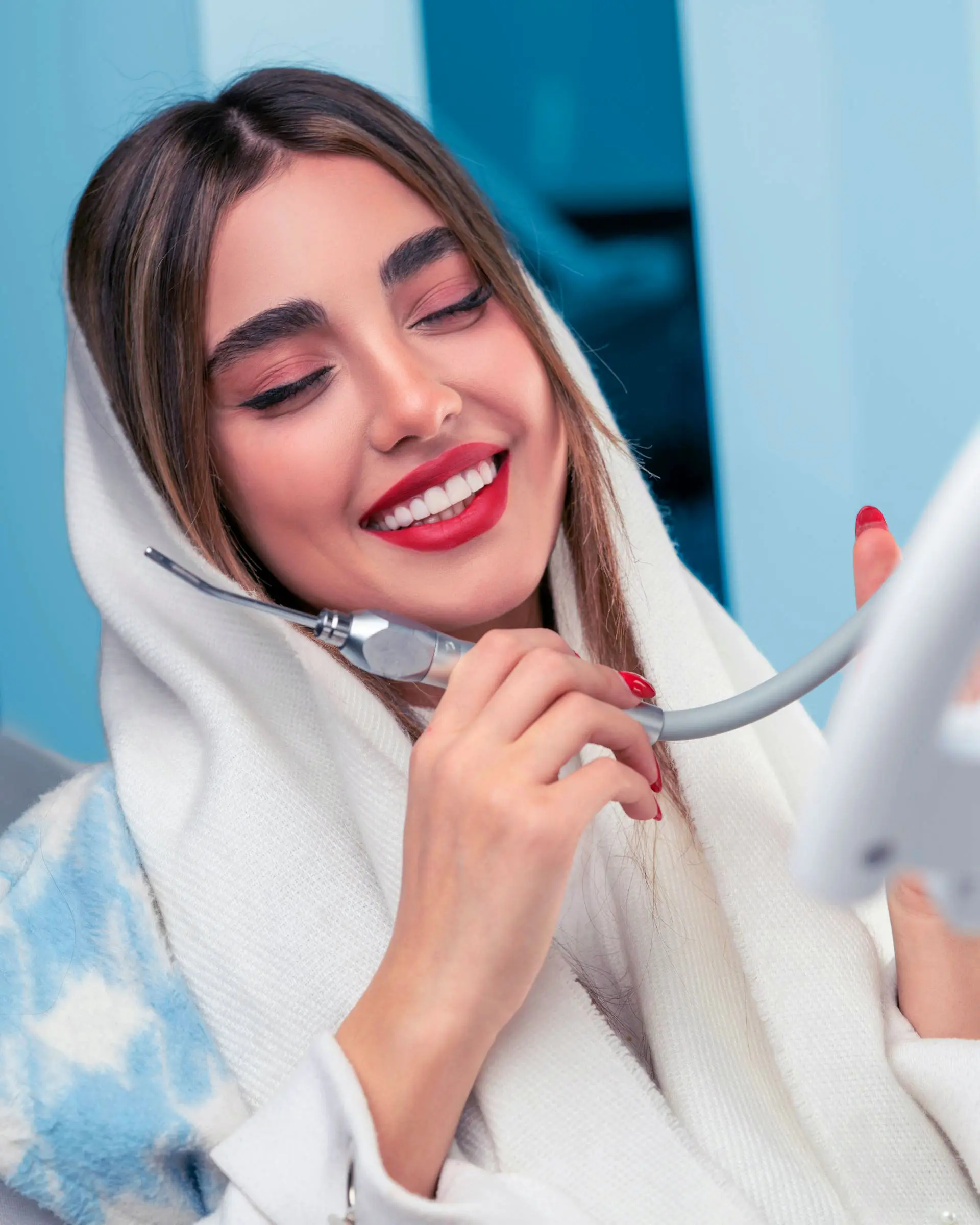 A smiling woman in a white hijab holds a device while smiling seated in a dentist chair against a blue background.