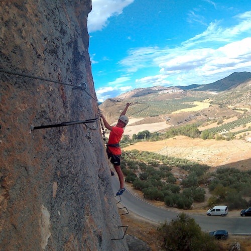 一位穿着红色衬衫的攀岩者正攀爬着一个陡峭的岩壁,岩壁上有金属梯级,俯瞰着一片丘陵起伏和道路蜿蜒的风景如画的景色。