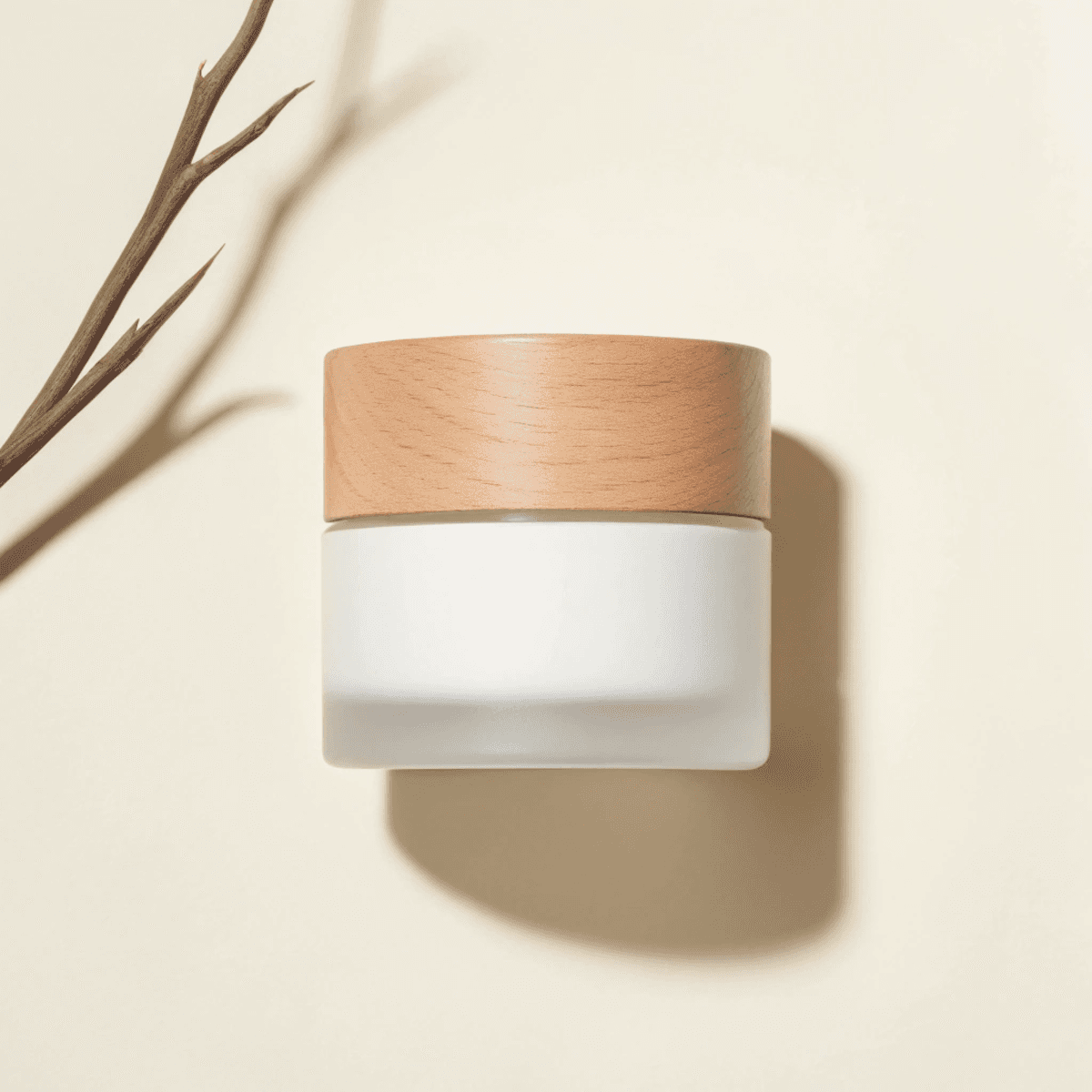 A minimalist skincare cream jar with a wooden lid is set against a neutral beige background, accompanied by a delicate twig, reflecting a natural and eco-friendly aesthetic.