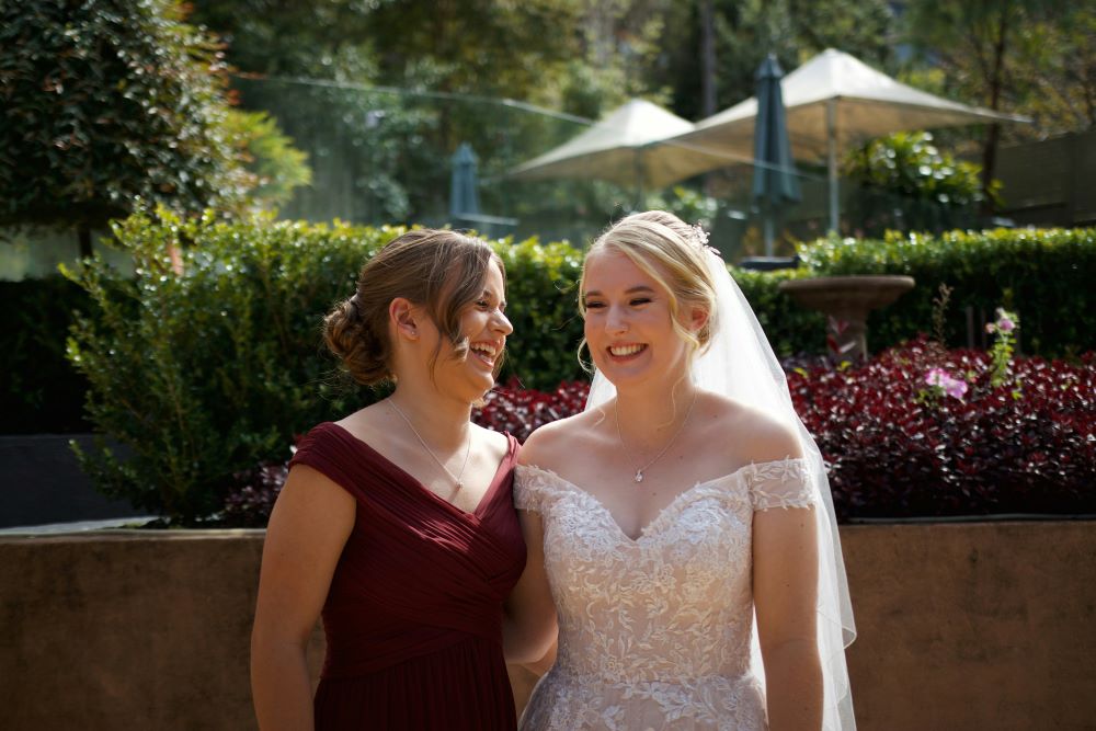Lachende Braut mit ihrer Schwester an ihrem Hochzeitstag: Die Braut trägt ein elegantes Spitzen-Hochzeitskleid mit Off-Shoulder-Ärmeln und einen langen Schleier, während ihre Schwester in einem weinroten Kleid an ihrer Seite steht. Beide lachen herzlich in einem sonnigen Garten mit grünen Pflanzen und Sonnenschirmen im Hintergrund. Maria Wiethaler freie Rednerin und Sängerin für Hochzeiten.
