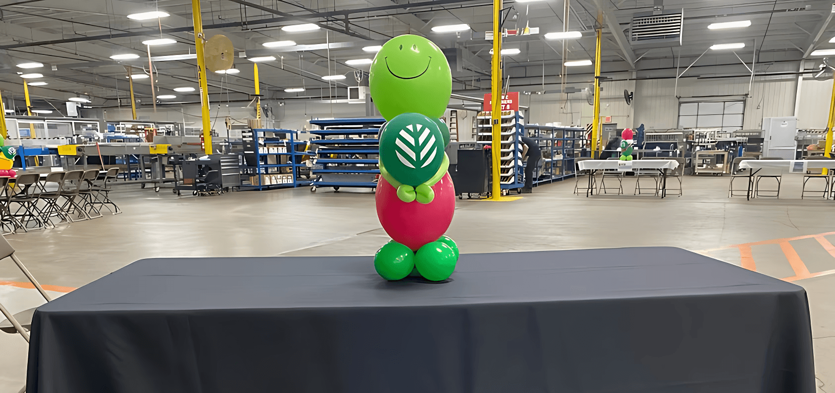 Colorful balloon centerpiece with red, green, and black balloons placed on a table in a large event hall by Be Bold Balloons